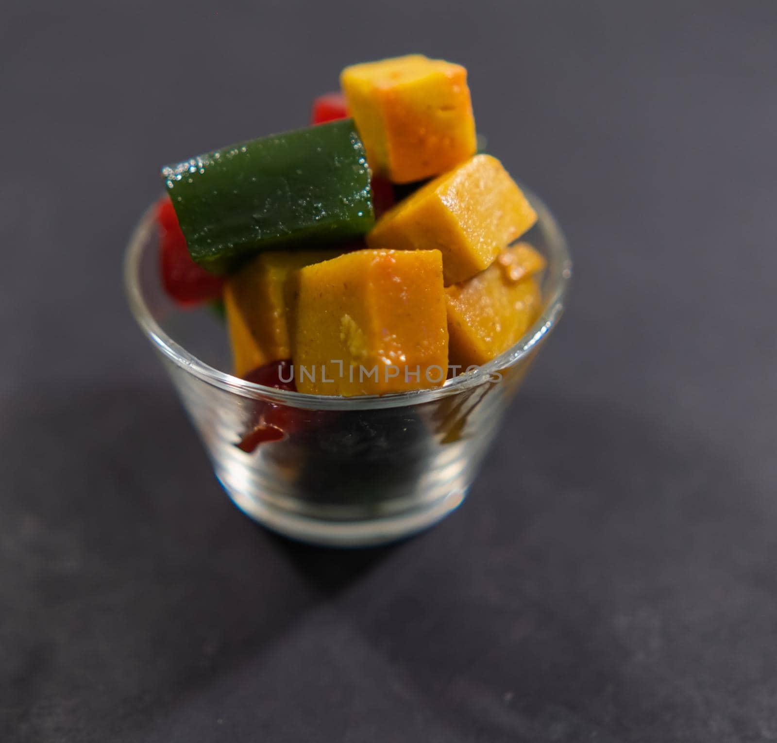 Close-up of glass of red, yellow, and green fruit paste slices on black surface. Authentic colorful and natural Mexican candy cubes in glass cup. Tasty traditional snacks