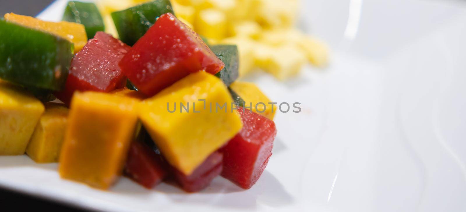 Extreme close-up of red, yellow, and green fruit paste cubes and diced cheese on white plate. Porcelain plate of fresh yellow cheese slices and traditional fruit candy. Sweet and healthy snacks