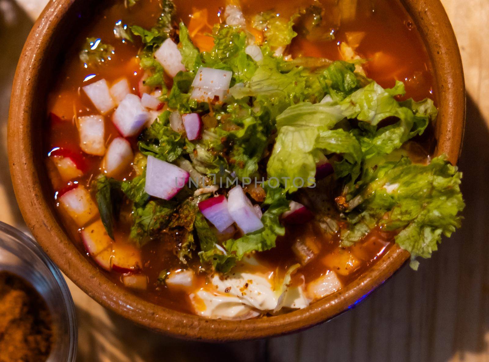 Top view of clay bowl of delicious and traditional red pozole on wooden table. Authentic Hispanic pork stew in handmade bowl with chopped radish and lettuce on top. Traditional Mexican cuisine