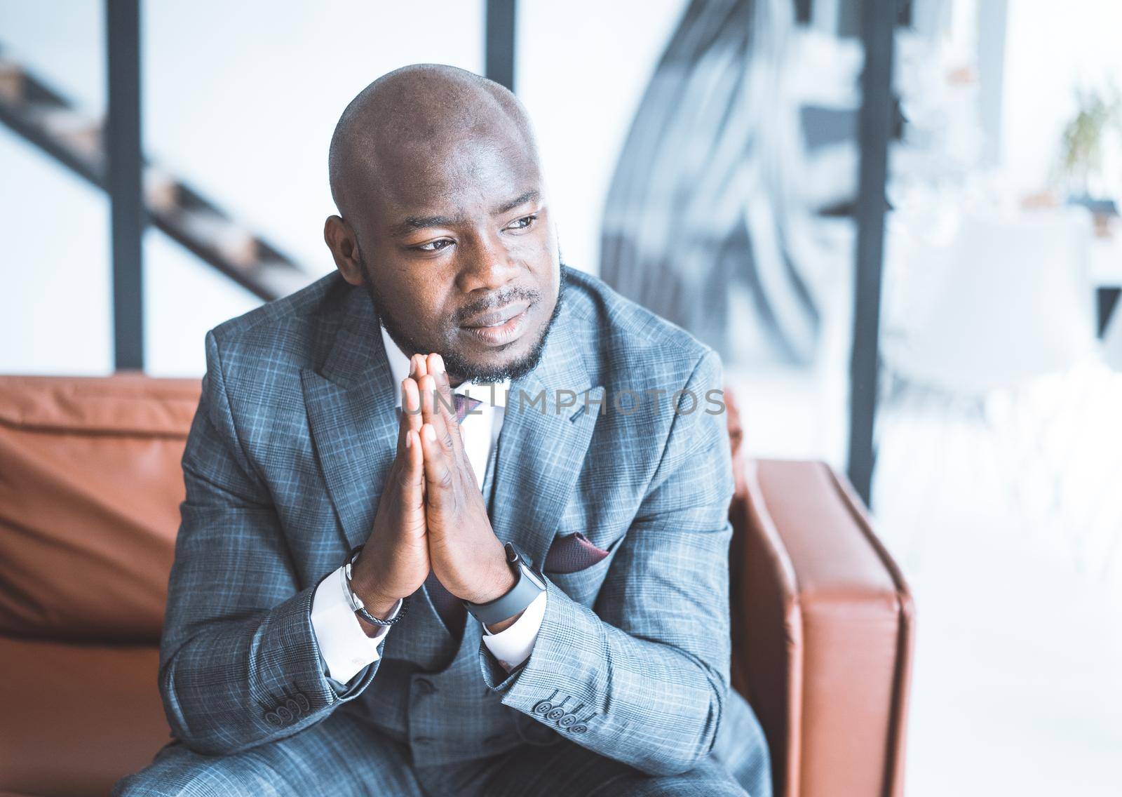 Serious Lawyer with Piercing Gaze in Formal Suit Sits on the Couch in his Office in a Futuristic Style. Male Model Puts his Palms of the Hands Together and Posing in Minimalism Light Studio. Close-up Portrait. Copy Space. High quality photo