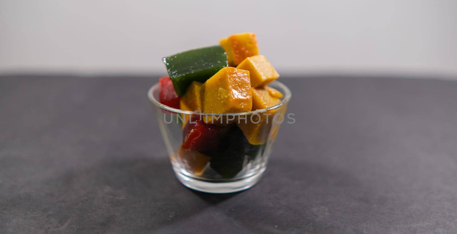 Wide view of glass of red, yellow, and green fruit paste slices above black and white background. Close-up of authentic natural Mexican candy cubes in cup above dark surface. Tasty traditional snacks
