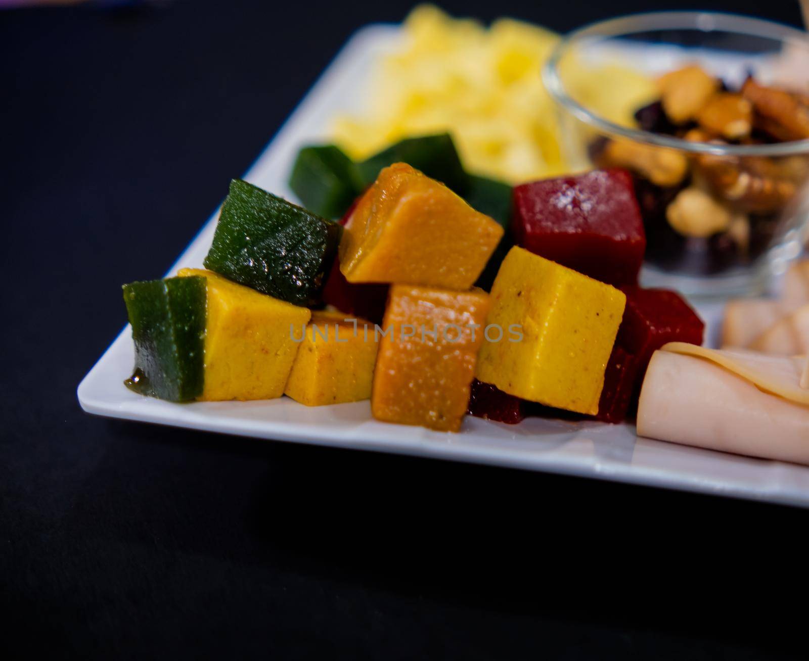 Close-up of diced fruit paste, turkey ham rolls, cheddar cheese cubes, and glass of walnuts on square white plate. Turkey meat, colorful Mexican candy, and nuts above black surface. Healthy snacks