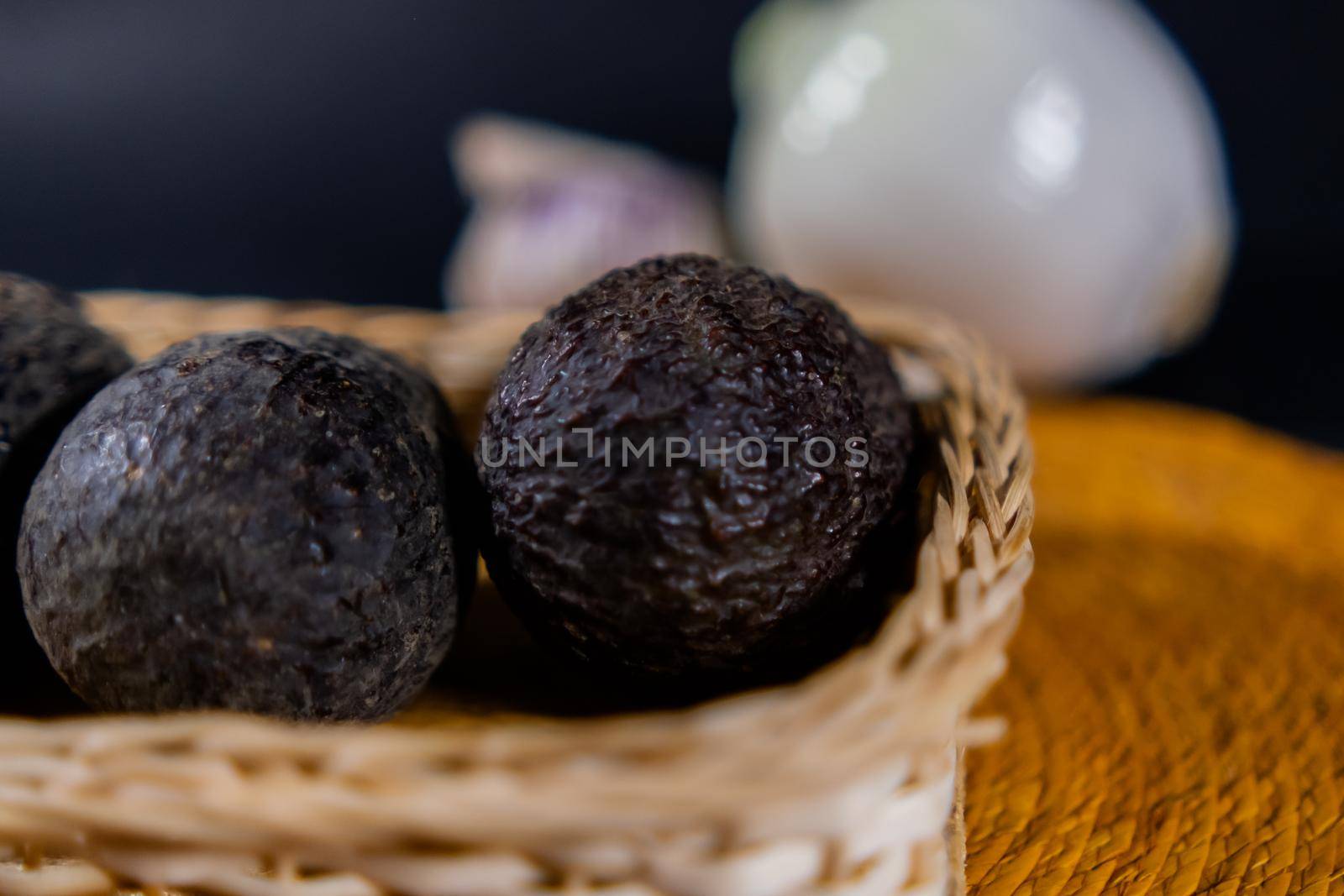 Close-up of three avocados in traditional wooden and palm basket with blurry onion and garlic as background. Fresh black vegetables in handmade basket. Authentic healthy food preparation