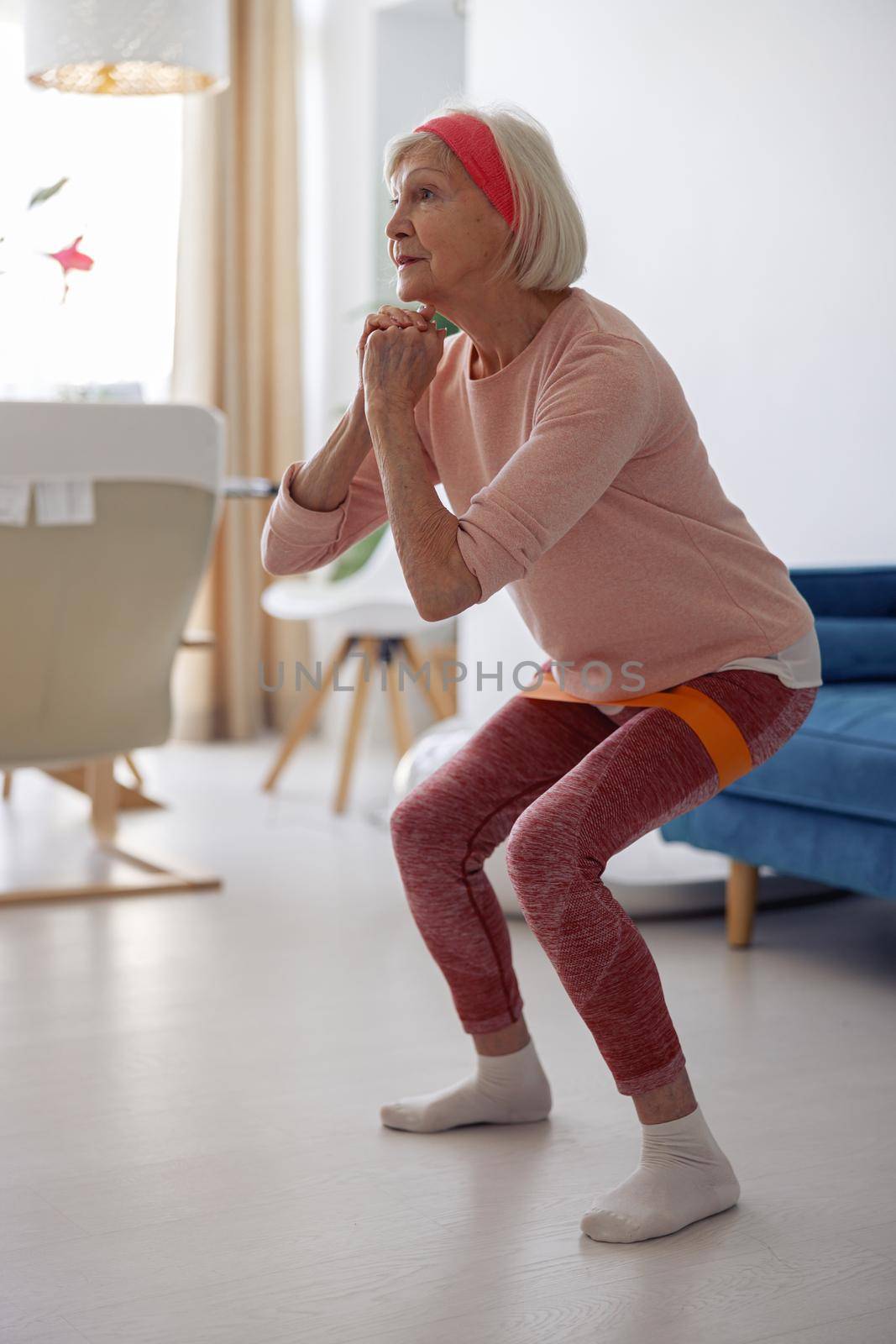 Active elderly lady working out while doing squats with elastic band on blurred interior of her living room