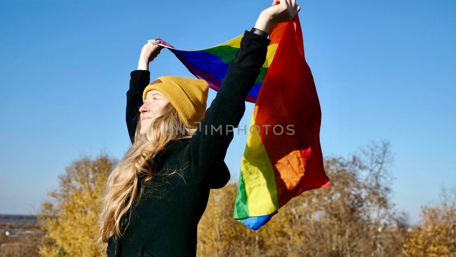 Portrait of a young, Caucasian, blonde, girl with acne holding a rainbow LGBT flag in autumn. Woman in yellow hat and black coat on a sunny day