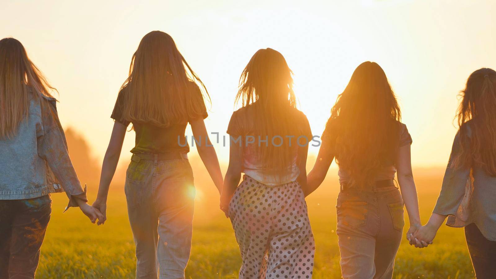 A group of girls walk towards the sun at sunset holding hands