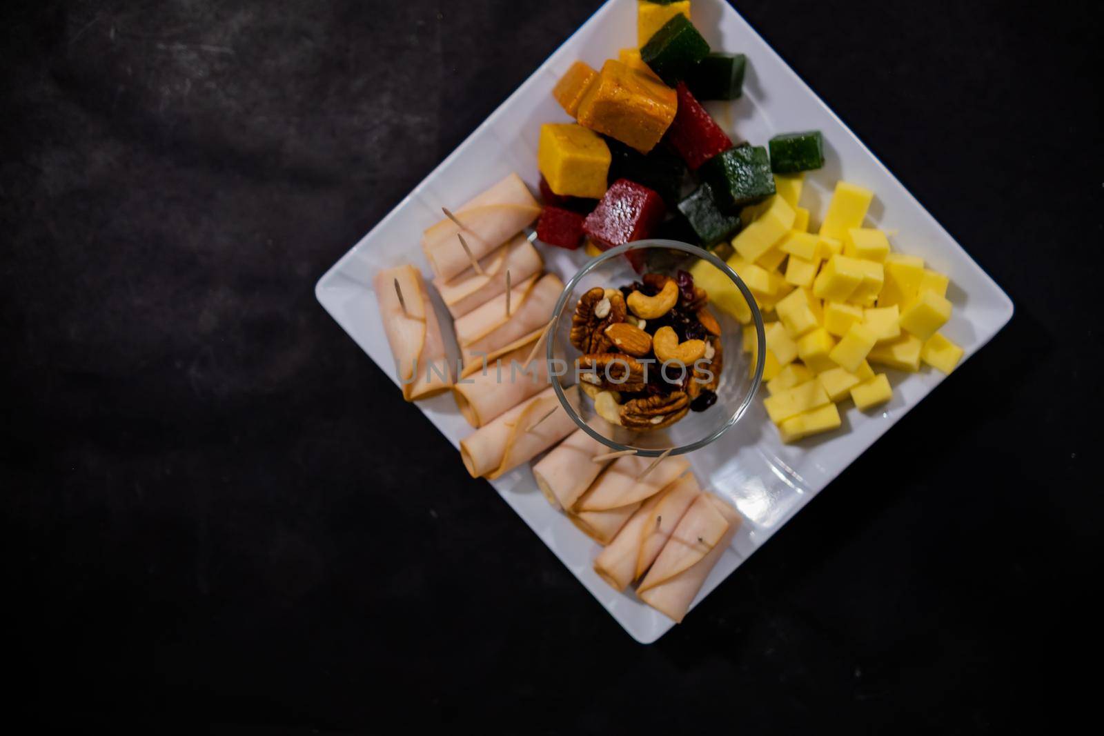 Top view of turkey ham rolls, diced fruit paste, cheddar cheese cubes, and glass of walnuts on square white plate. Turkey meat, colorful Mexican candy, and nuts above black surface. Healthy snacks