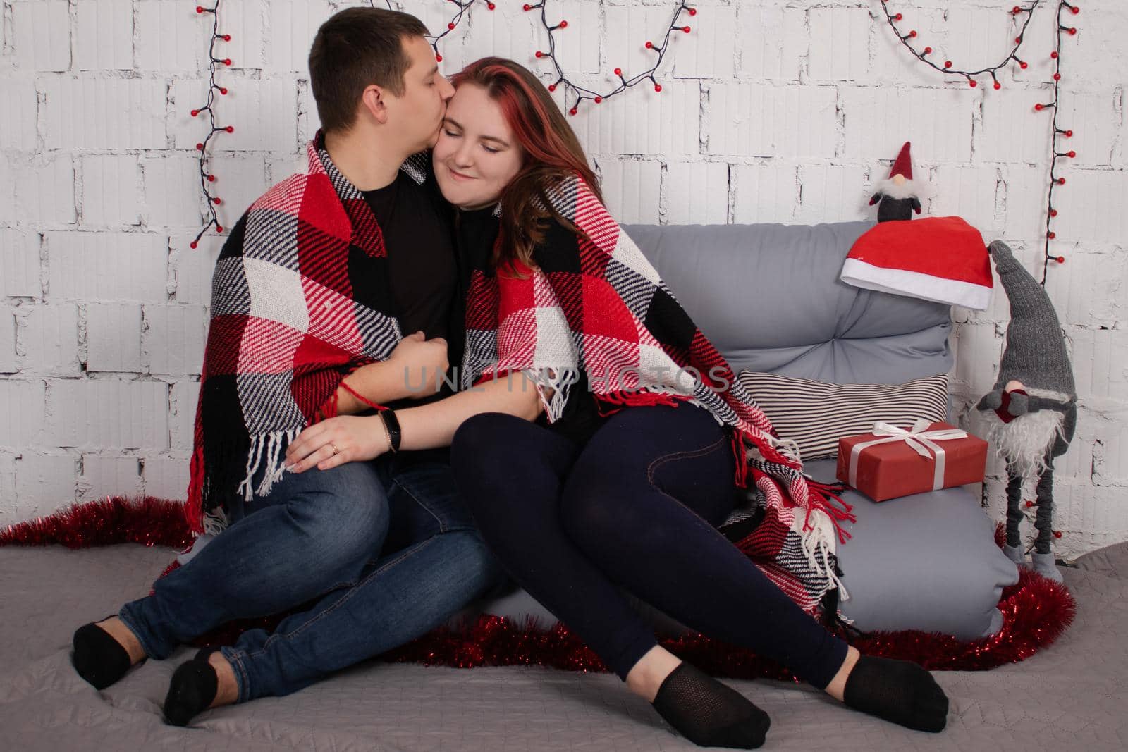 happy couple hugging on grey couch covered in red checkered plaid-blanket near red christmas decor. cozy young family.