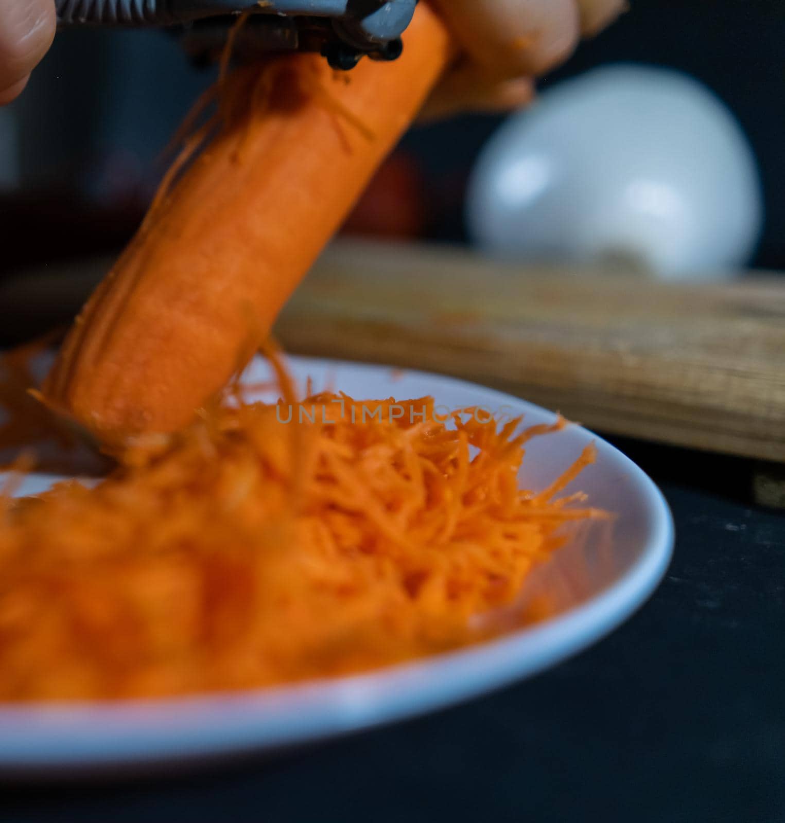 Close-up of hands peeling carrot with julienne peeler above white plate with blurry vegetables as background. Person making small pile of grated orange vegetables. Healthy food preparation