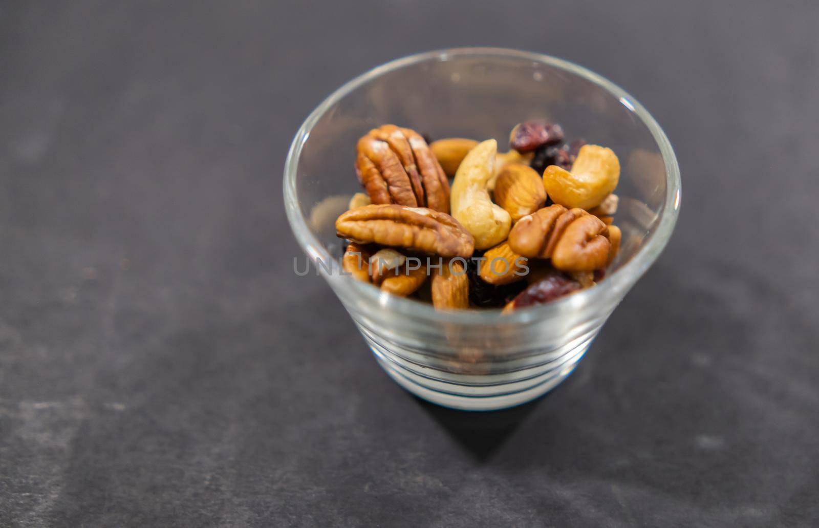 Close-up of small glass of walnuts, almonds, Indian nuts, and dry cranberries on black surface. Fresh natural nuts in glass cup above dark background. Tasty traditional snacks