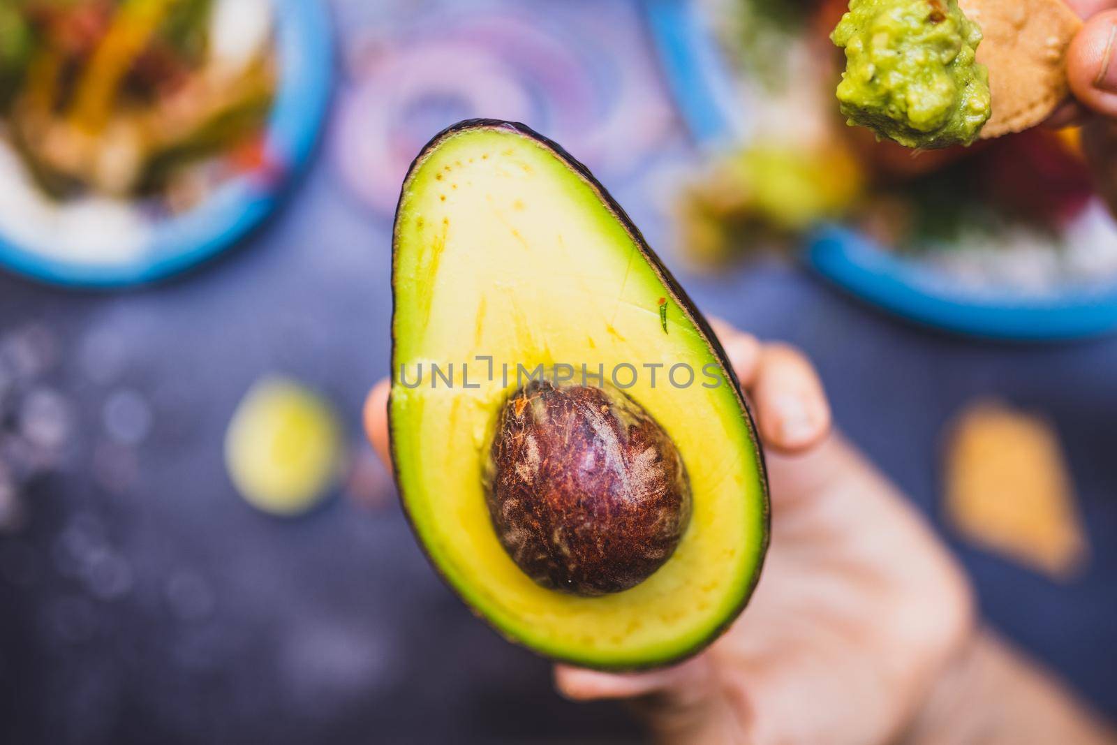 Close-up of hand holding an avocado half with pit above blurry background. Top view of person holding traditional Mexican fruit and tortilla chip with guacamole above blurry table. Authentic food