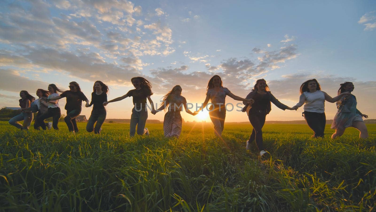 Eleven cheerful girls run to the meeting across the field in the summer, holding hands