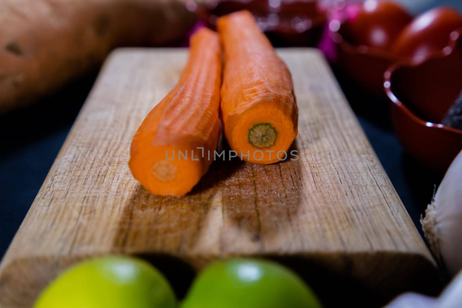 Close-up of two peeled carrots on wooden cutting board surrounded by vegetables in clay pots. Fresh and thin carrots, limes, onion, avocado, and more above black surface. Healthy food preparation