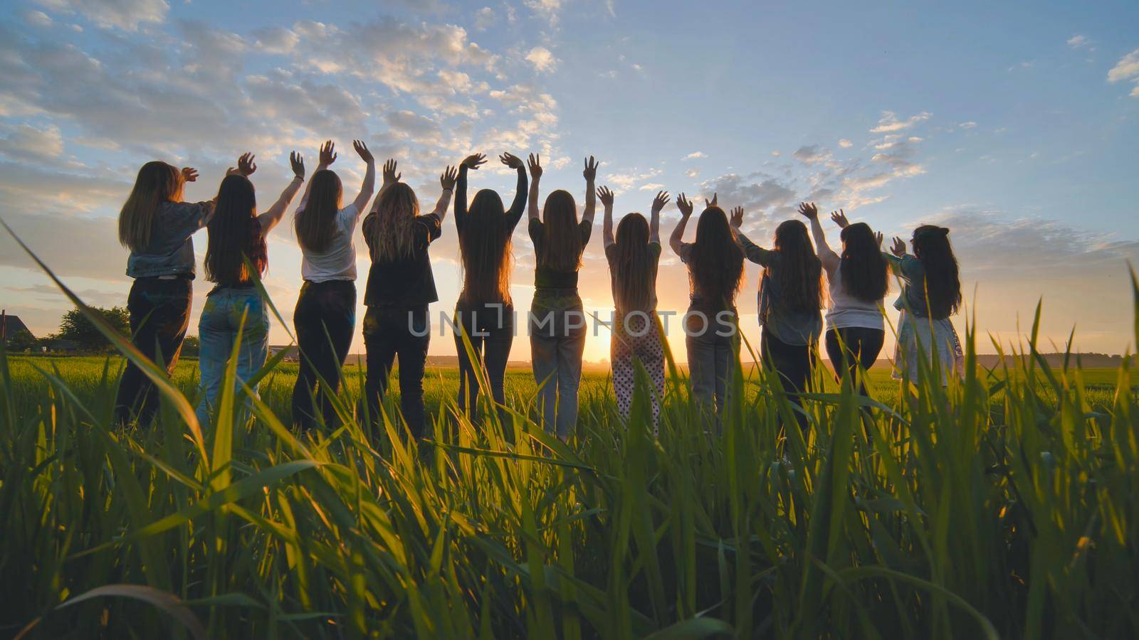 Silhouette of friends of 11 girls waving their hands at sunset in the field. by DovidPro