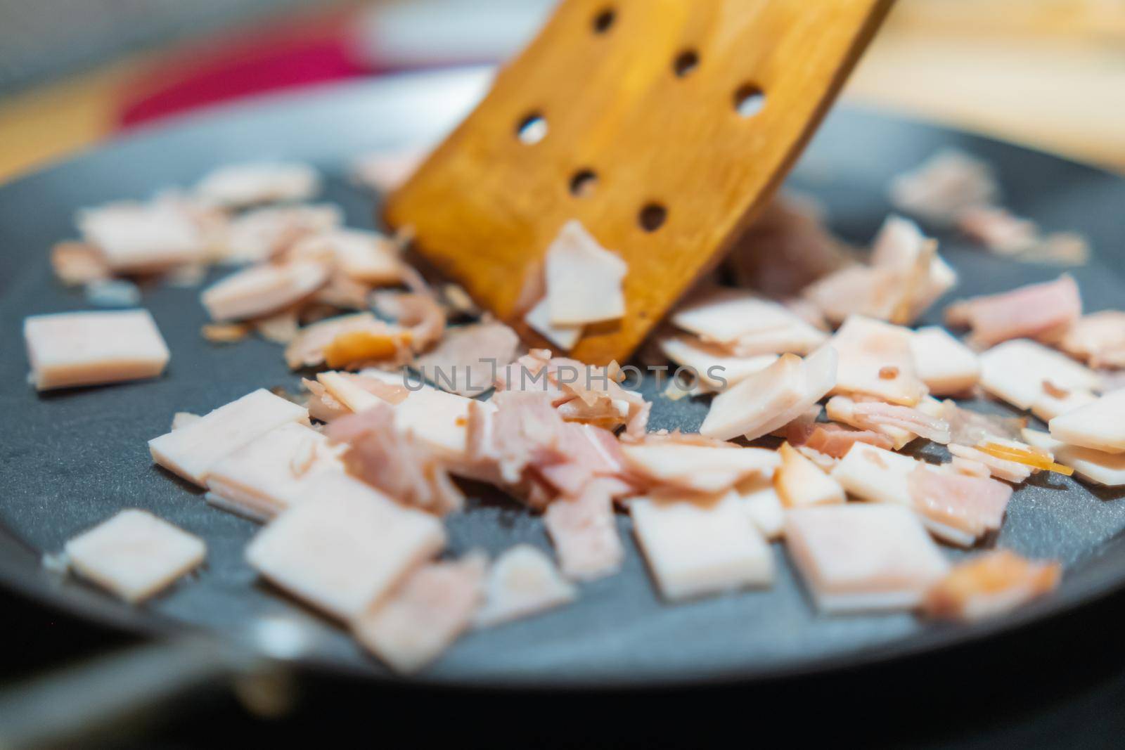 Close-up of wooden spoon moving chopped turkey ham above round griddle. Cooking thin turkey breast meat cut into squares on metal surface. Healthy meal preparation