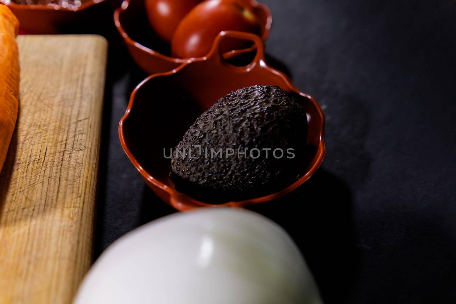 Close-up of two tomatoes, onion, and avocado in traditional clay pots above black surface. Fresh vegetables in handmade clay bowls around wooden cutting board. Authentic food preparation