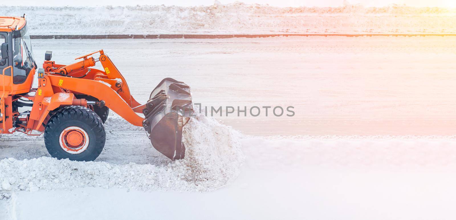 Big orange tractor cleans up snow from the road and loads it into the truck. Cleaning and cleaning of roads in the city from snow in winter in the rays of sunlight