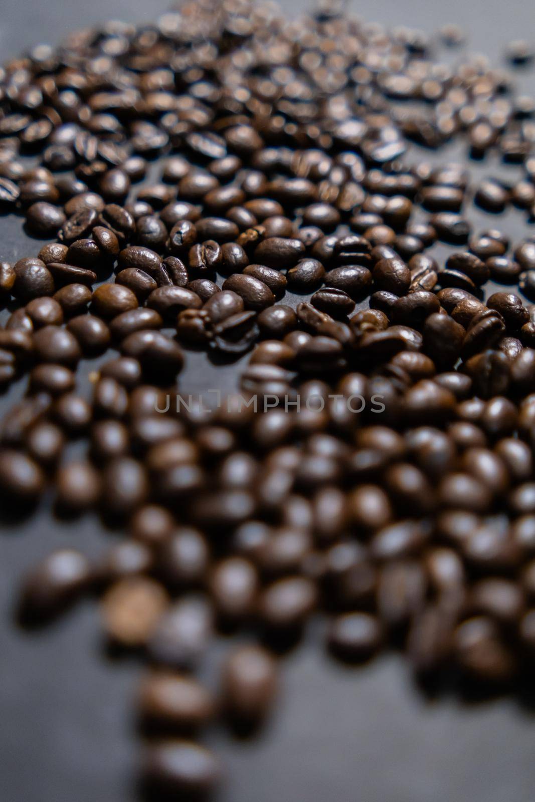 Close-up of small pile of roasted coffee beans on dark gray surface. Group of coffee grains scattered above dark color background. Caffeine and drink preparation