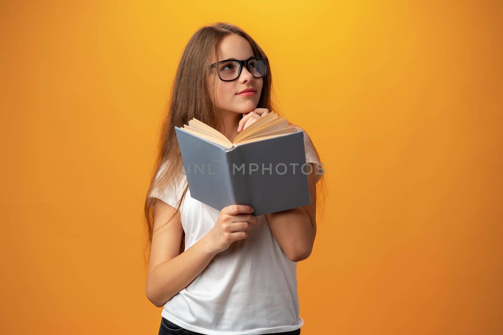 Smart teen girl in glasses holding book against yellow background, close up
