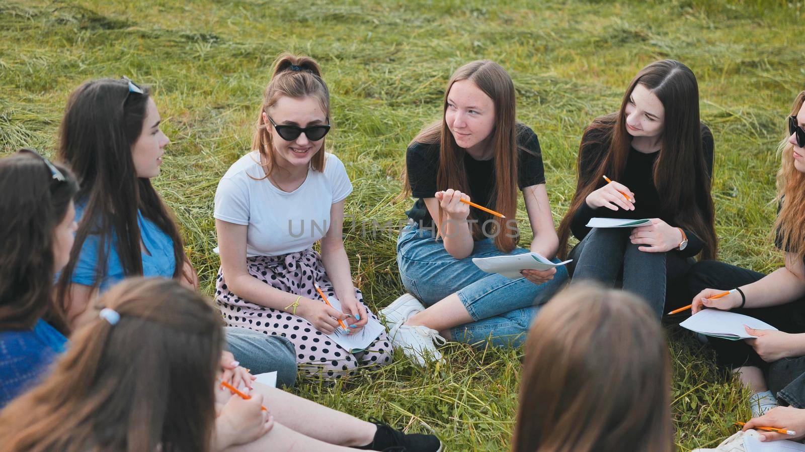 A group of female students are sitting in a circle on a meadow for collective work with notebooks. by DovidPro