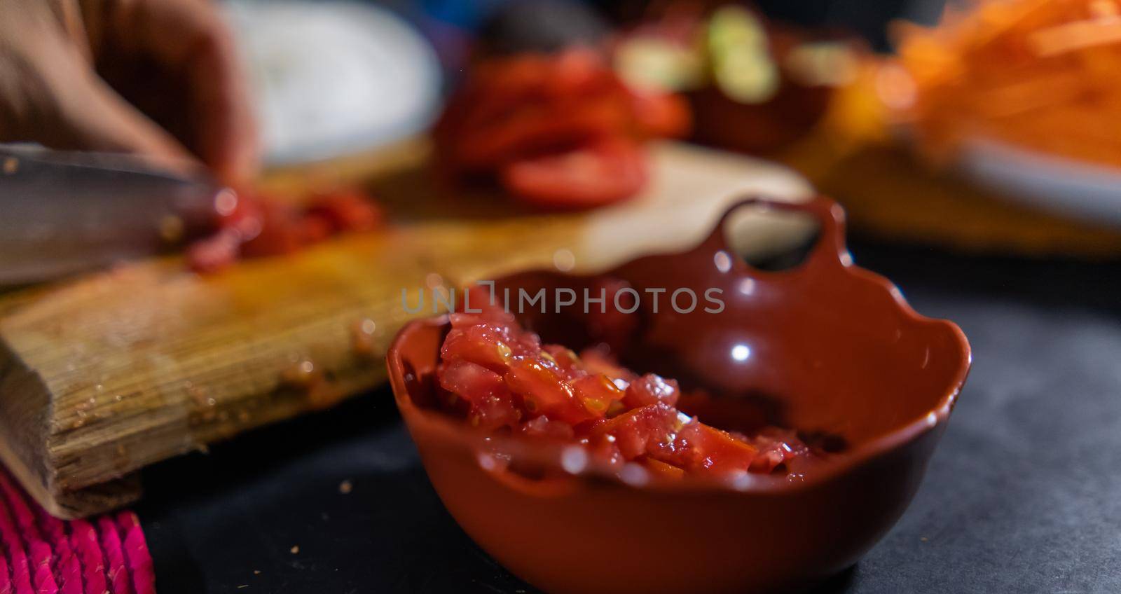 Close-up of chopped tomato in small clay pot with hands slicing more tomato as background. Person cutting fresh red vegetable with big knife on wooden cutting board. Healthy meal preparation