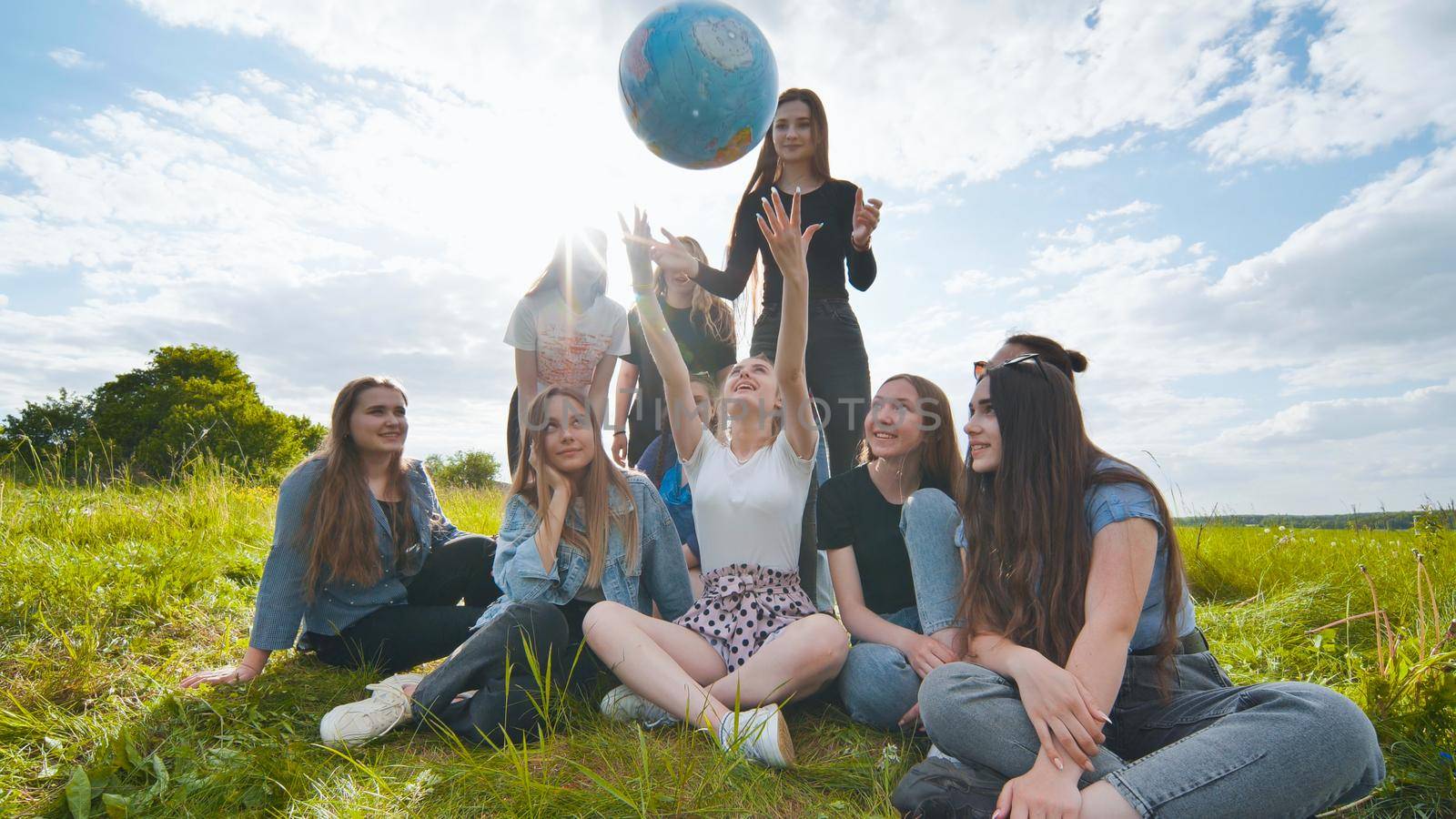 A group of girls students sit in the meadow and toss the world globe up. by DovidPro