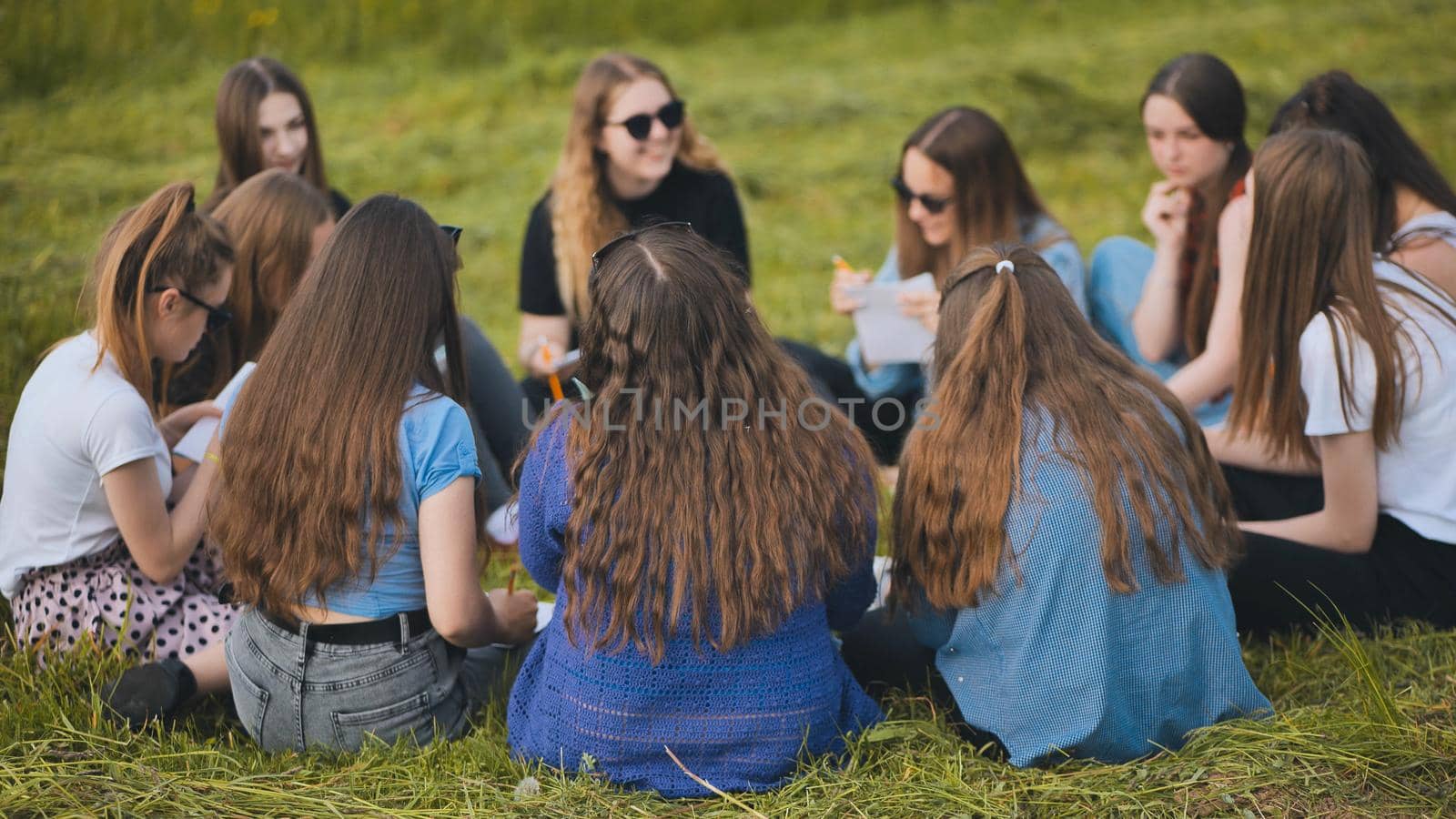 A group of female students are sitting in a circle on a meadow for collective work with notebooks