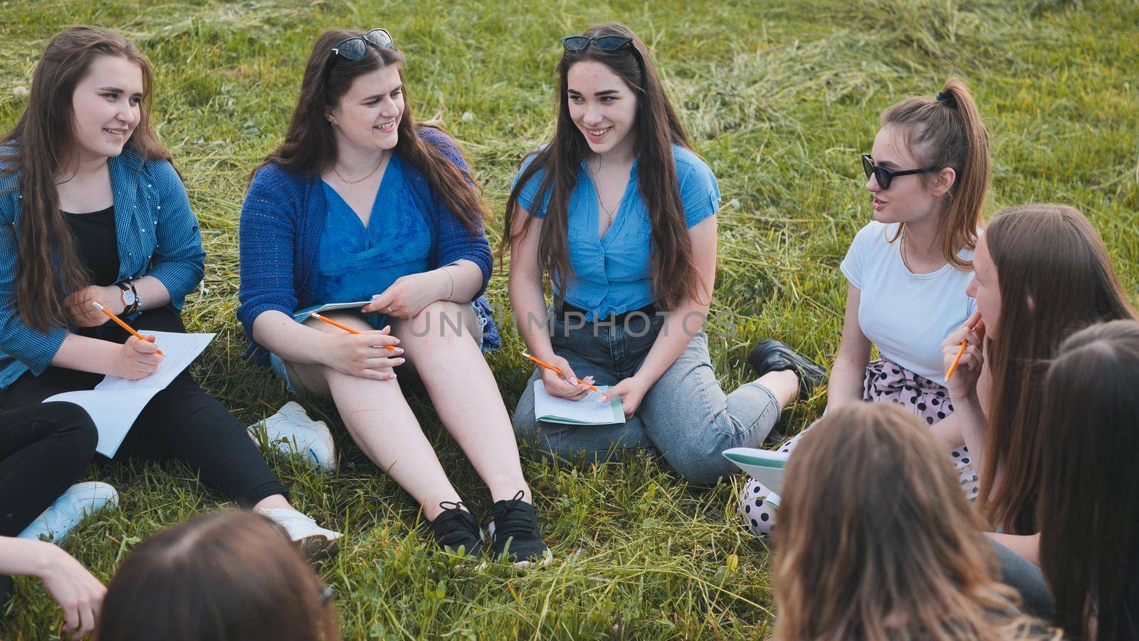 A group of female students are sitting in a circle on a meadow for collective work with notebooks