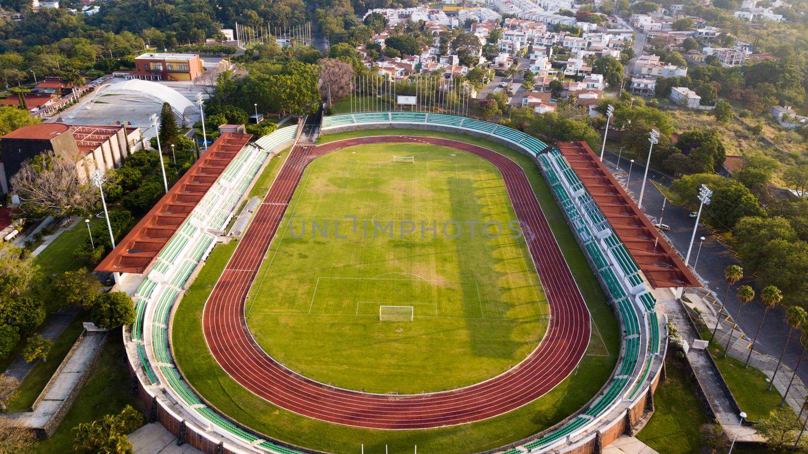 Beautiful aerial view of running track around soccer field with small town and trees as background. Peaceful view of small college stadium surrounded by houses and green foliage. Athletics and sports