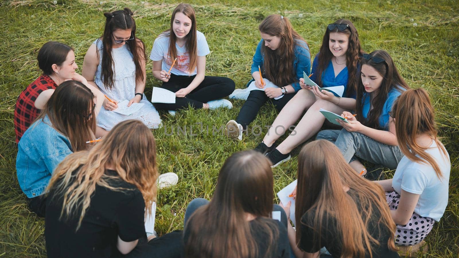 A group of female students are sitting in a circle on a meadow for collective work with notebooks