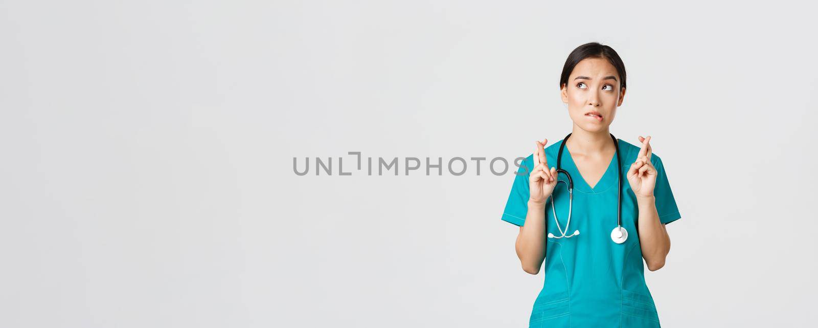 Covid-19, healthcare workers, pandemic concept. Hopeful nervous asian female intern in scrubs waiting for news, doctor cross fingers and biting lip as looking upper left corner, praying by Benzoix