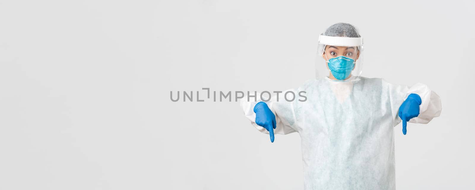 Covid-19, coronavirus disease, healthcare workers concept. Surprised and excited female doctor, physician in personal protective equipment pointing fingers down, standing white background.