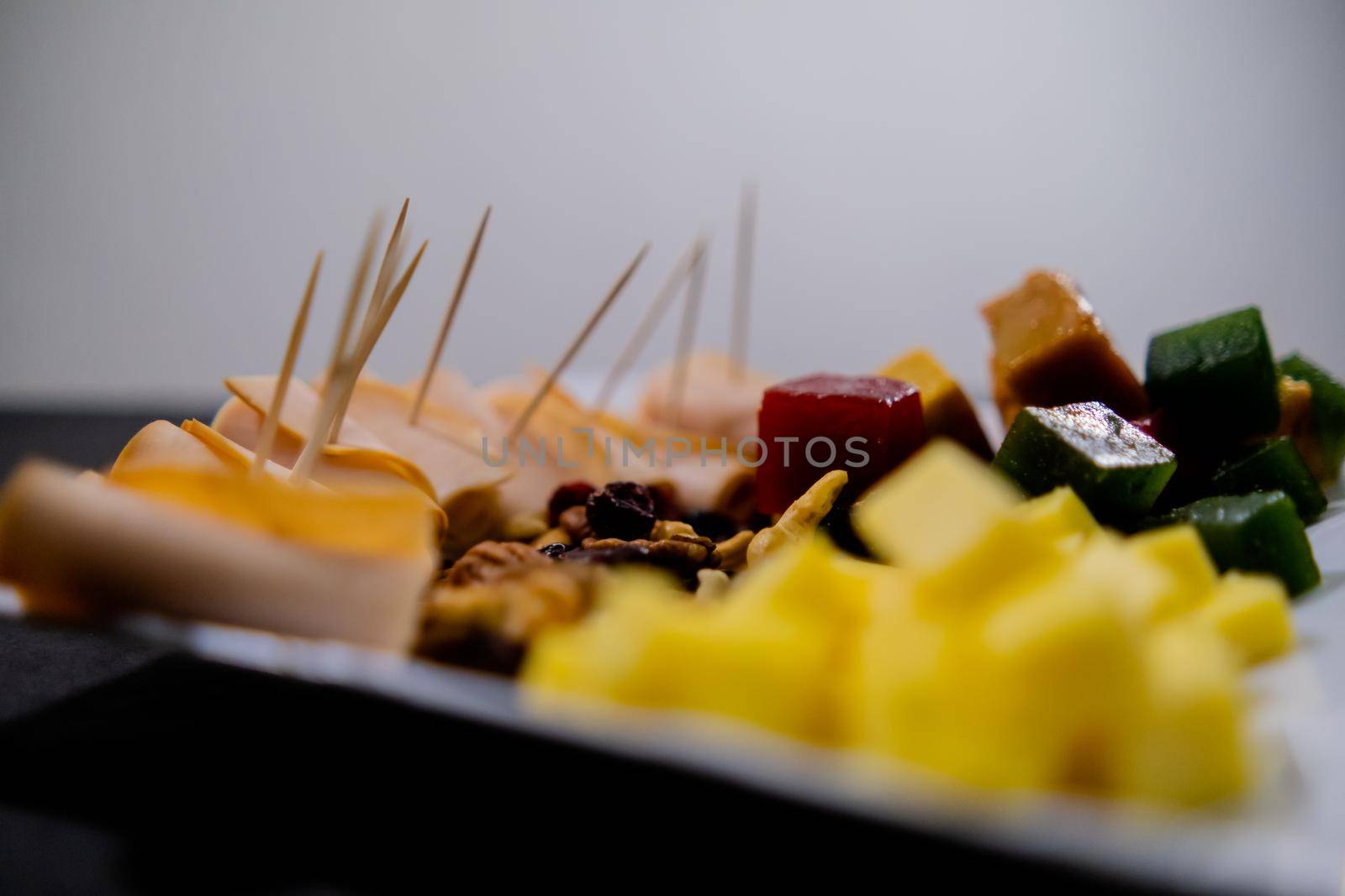 Close-up of turkey ham rolls, diced fruit paste, cheddar cheese cubes, and walnuts on square white plate. Turkey meat, colorful Mexican candy, and nuts with white background. Healthy snacks