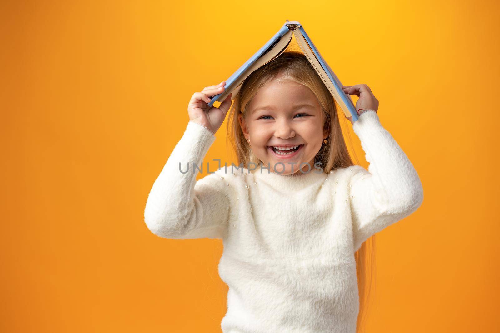 Little girl holding book on her head and looking at camera over yellow background, close up