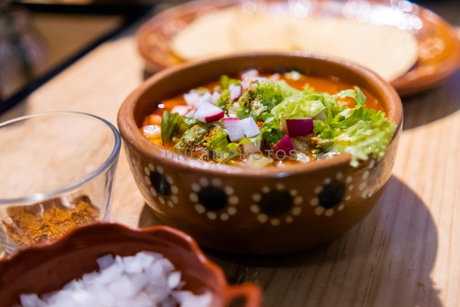 Close-up of clay bowl of delicious and traditional red pozole on wooden table. Authentic Hispanic pork stew in handmade bowl with chopped radish and lettuce on top. Traditional Mexican cuisine