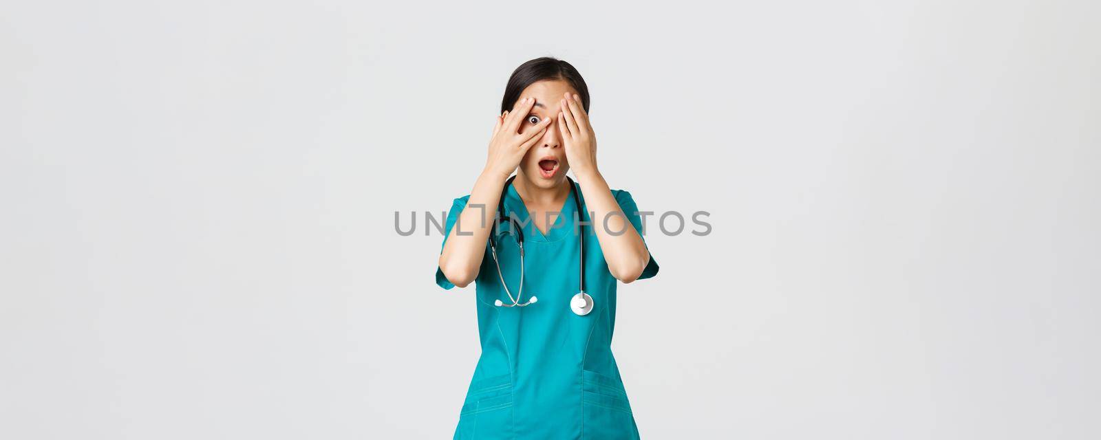 Covid-19, healthcare workers and preventing virus concept. Scared and shocked asian female nurse, doctor in scrubs witness something embarrassing or scary, cover eyes but peek through fingers.
