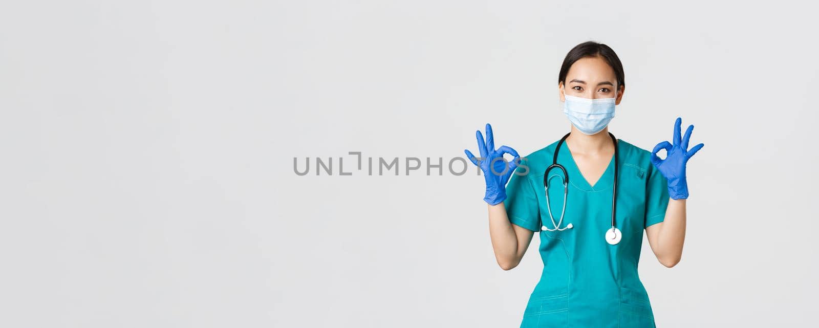 Covid-19, coronavirus disease, healthcare workers concept. Smiling professional and confident asian doctor, physician in medical mask, gloves and scrubs show okay gesture, guarantee quality service.