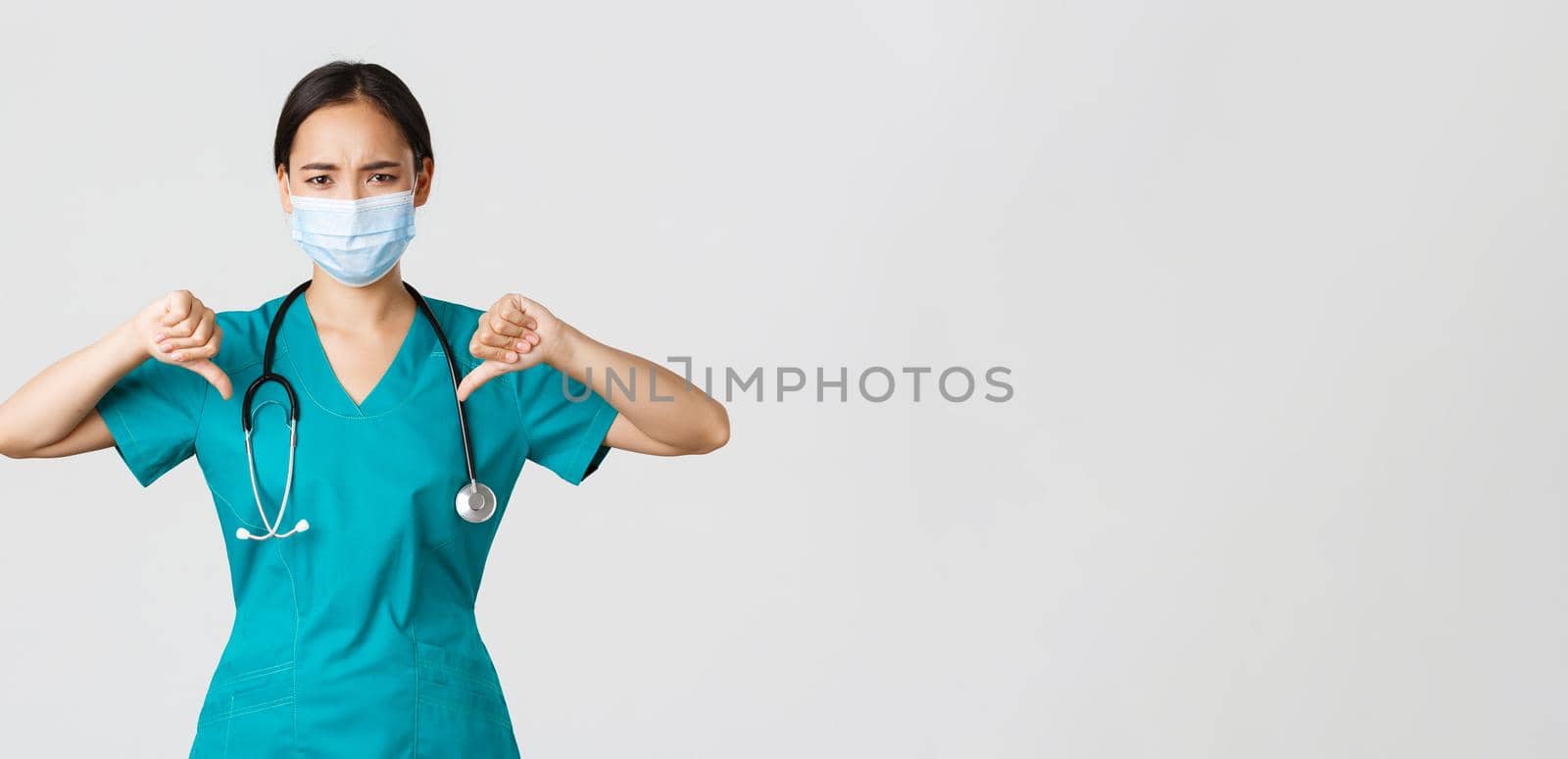 Covid-19, coronavirus disease, healthcare workers concept. Disappointed and upset asian female doctor in scrubs and medical mask showing thumbs-up, disagree or prohibit something, give advice by Benzoix