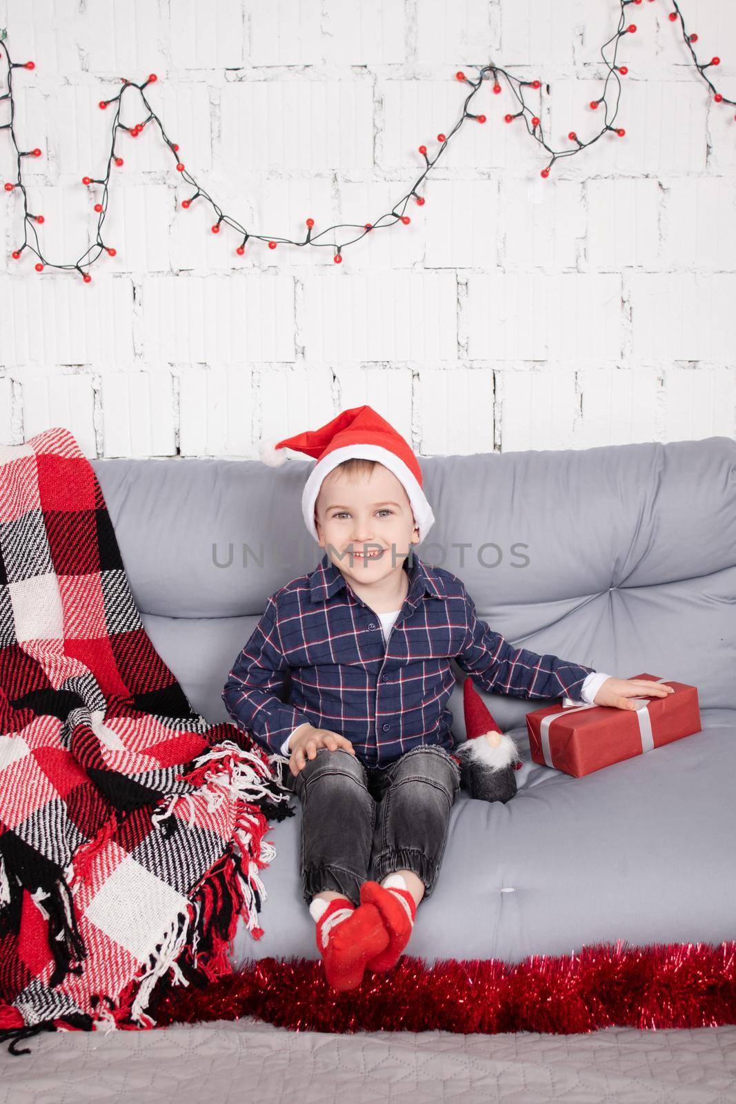 little boy in santa hat near christmas gnome toy, holding dwarf toy in hand in grey room. christmas decor and toys. kids playing by oliavesna