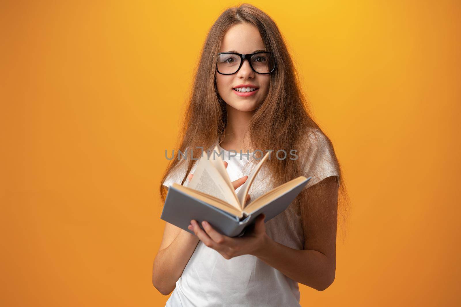 Smart teen girl in glasses holding book against yellow background, close up