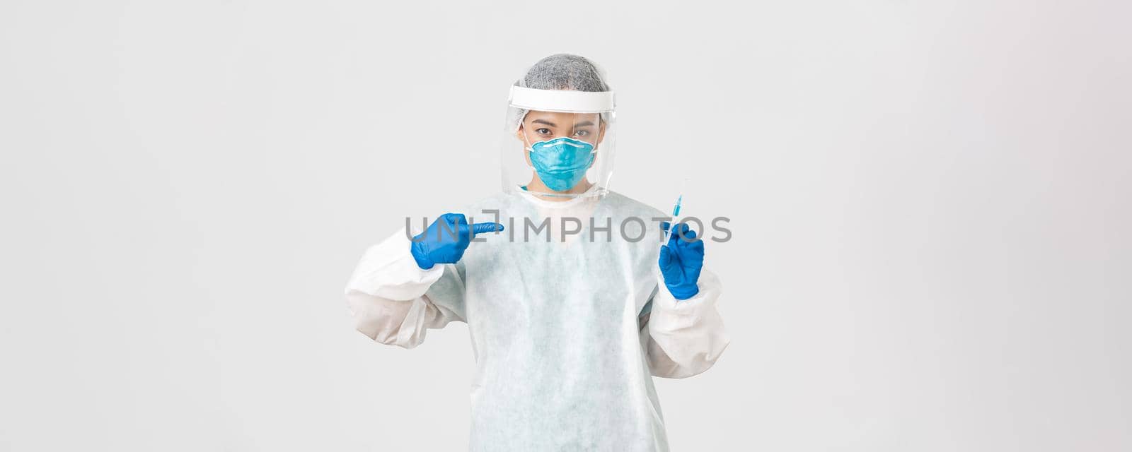 Covid-19, coronavirus disease, healthcare workers concept. Serious asian female doctor, physician in PPE personal protective equipment, pointing finger at syringe with vaccine, white background.