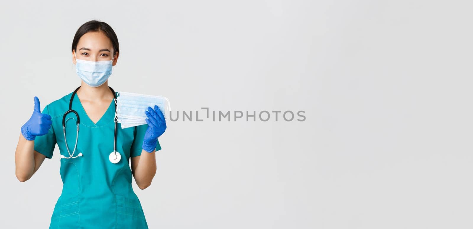 Covid-19, coronavirus disease, healthcare workers concept. Smiling asian doctor, nurse in scrubs and rubber gloves, showing thumbs-up and medical masks, white background by Benzoix