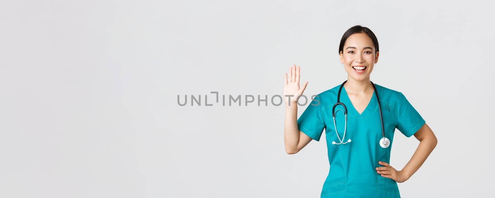 Covid-19, healthcare workers and preventing virus concept. Friendly-looking smiling asian female doctor, physician in scrubs waving hand to say hi, hello, greeting patient, nice to see you.