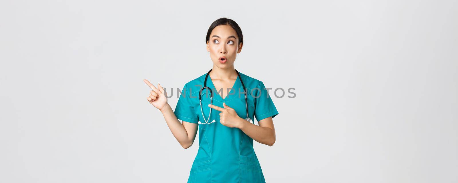 Covid-19, healthcare workers, pandemic concept. Intrigued and amazed beautiful asian female intern, nurse in scrubs or surgeon pointing and looking upper left corner, saying wow in amazement.