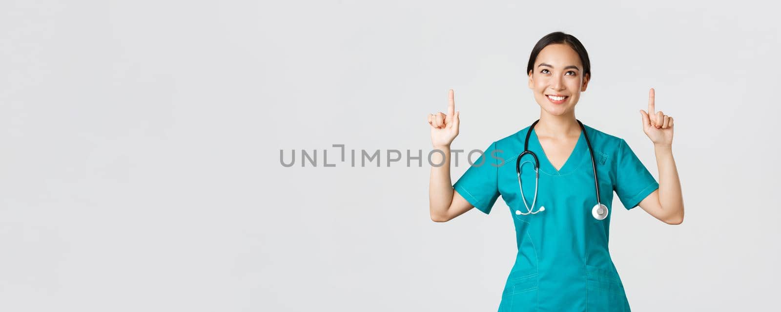 Covid-19, healthcare workers, pandemic concept. Smiling beautiful asian female nurse, doctor in scrubs pointing and looking up with satisfied grin, happy to show awesome promo offer, white background.