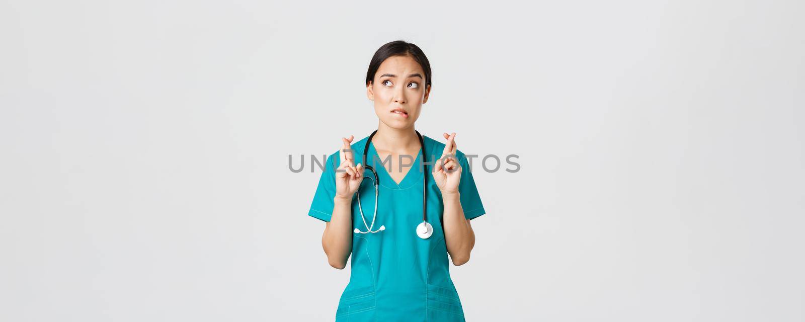 Covid-19, healthcare workers, pandemic concept. Hopeful nervous asian female intern in scrubs waiting for news, doctor cross fingers and biting lip as looking upper left corner, praying.