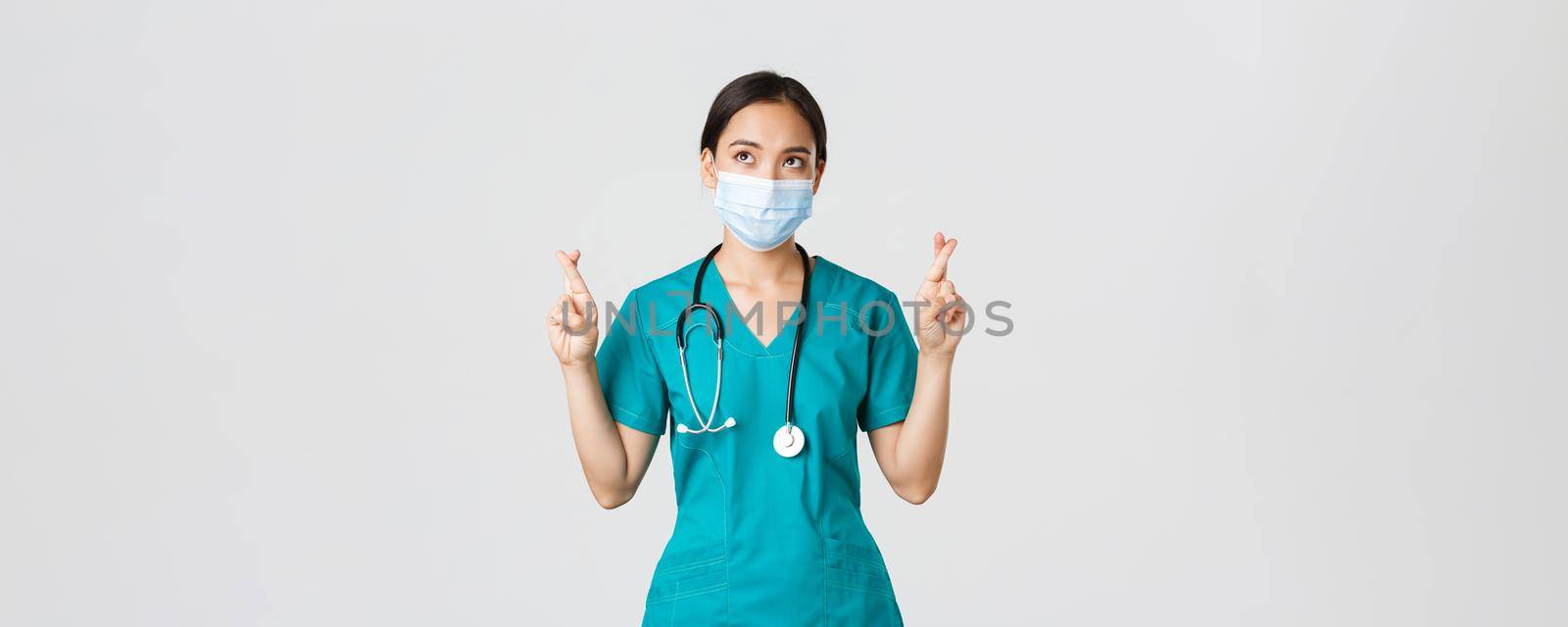 Covid-19, coronavirus disease, healthcare workers concept. Hopeful serious-looking asian doctor, female physician in medical mask and scrubs cross fingers, looking upper left corner by Benzoix