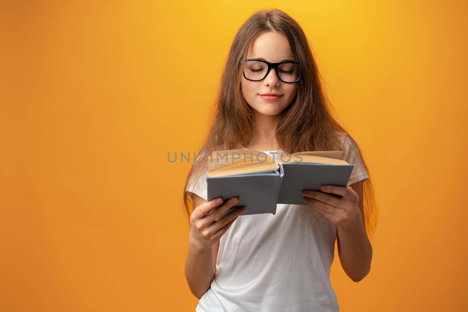 Smart teen girl in glasses holding book against yellow background, close up