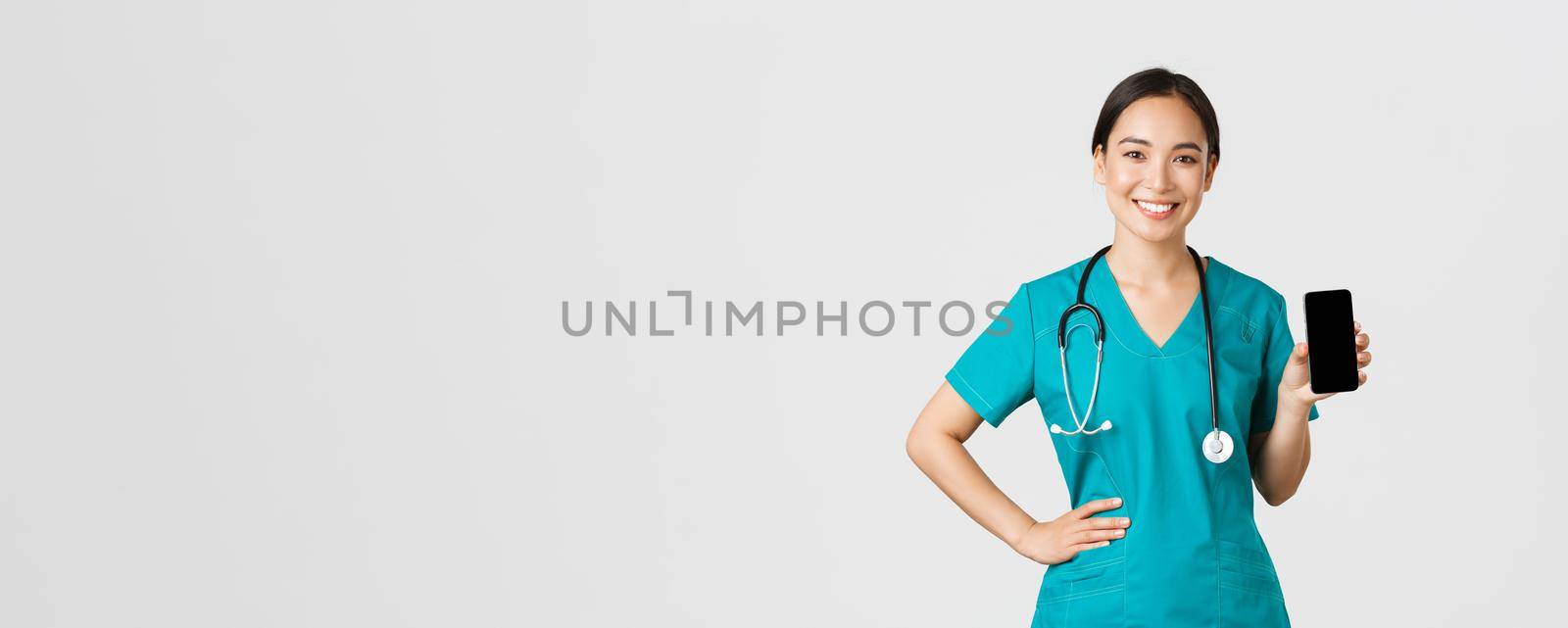 Covid-19, healthcare workers and online medicine concept. Professional female asian doctor, nurse in scrubs showing smartphone screen and smiling, showing internet appointment app, white background.