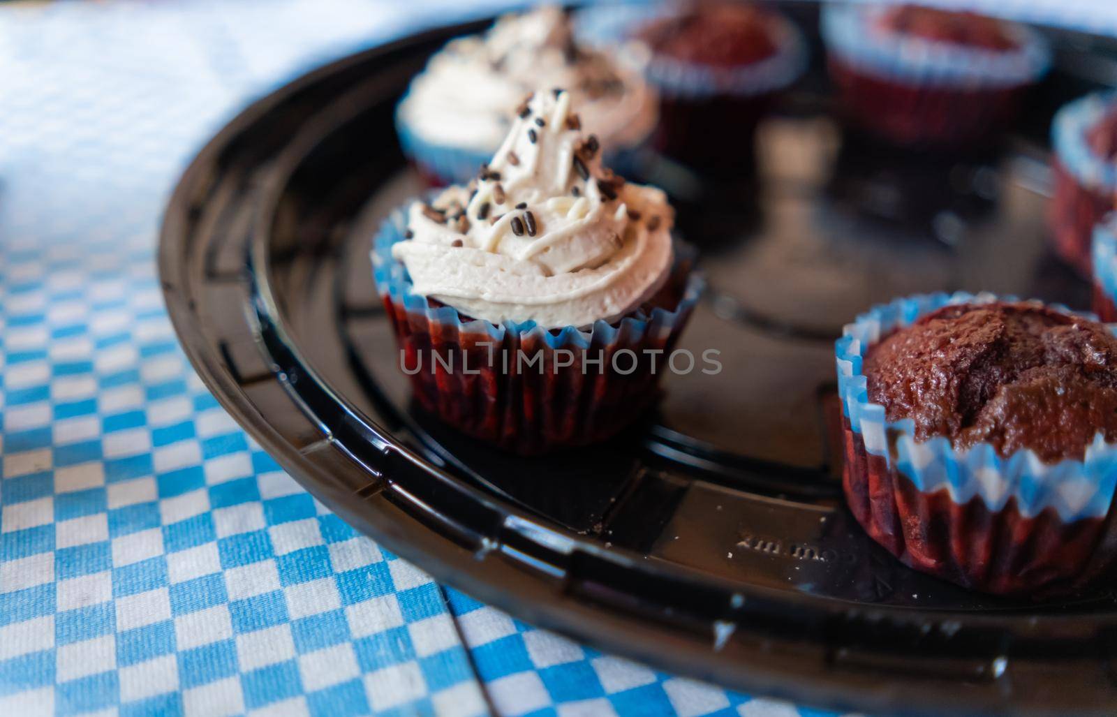Close-up of two delicious cupcakes and five chocolate muffins on round black plastic tray. Cute pastries with frosting and sprinkles above blue checkered tablecloth. Desserts and sweet food